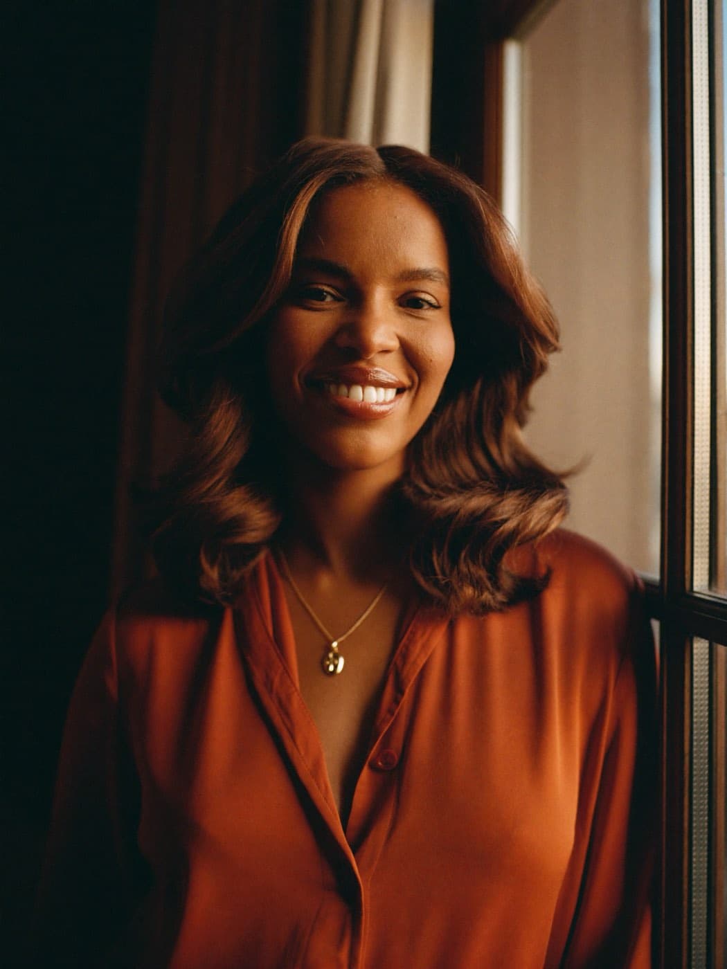 A woman in a terracotta blouse smiling softly by a tall window in golden-hour light