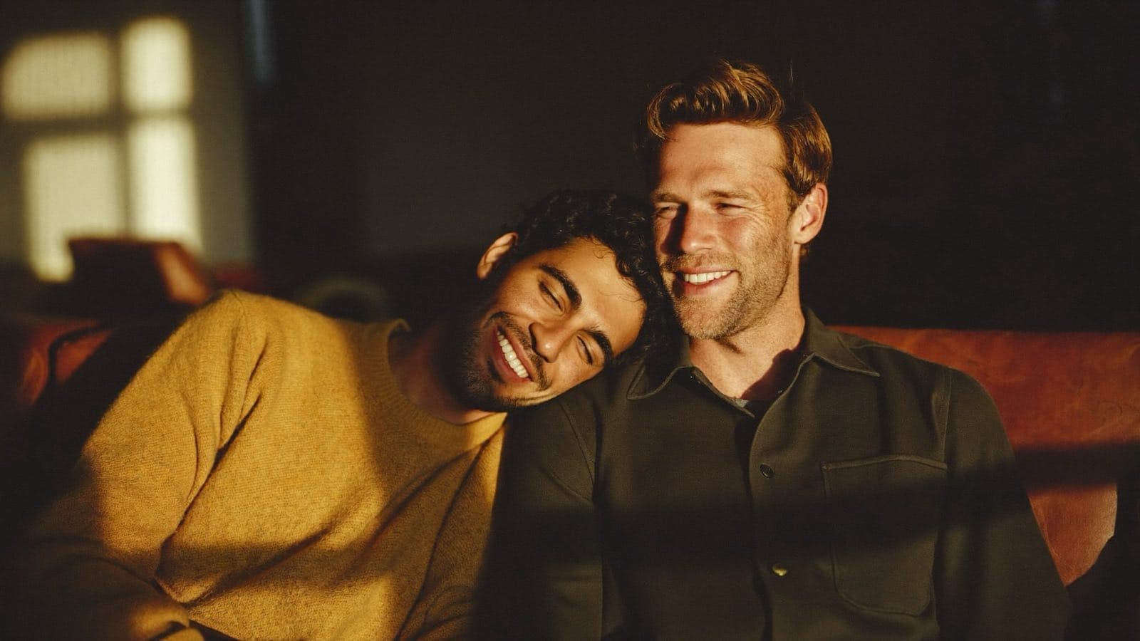 Two men laughing together on a sofa in golden afternoon light, one resting his head on the other's shoulder