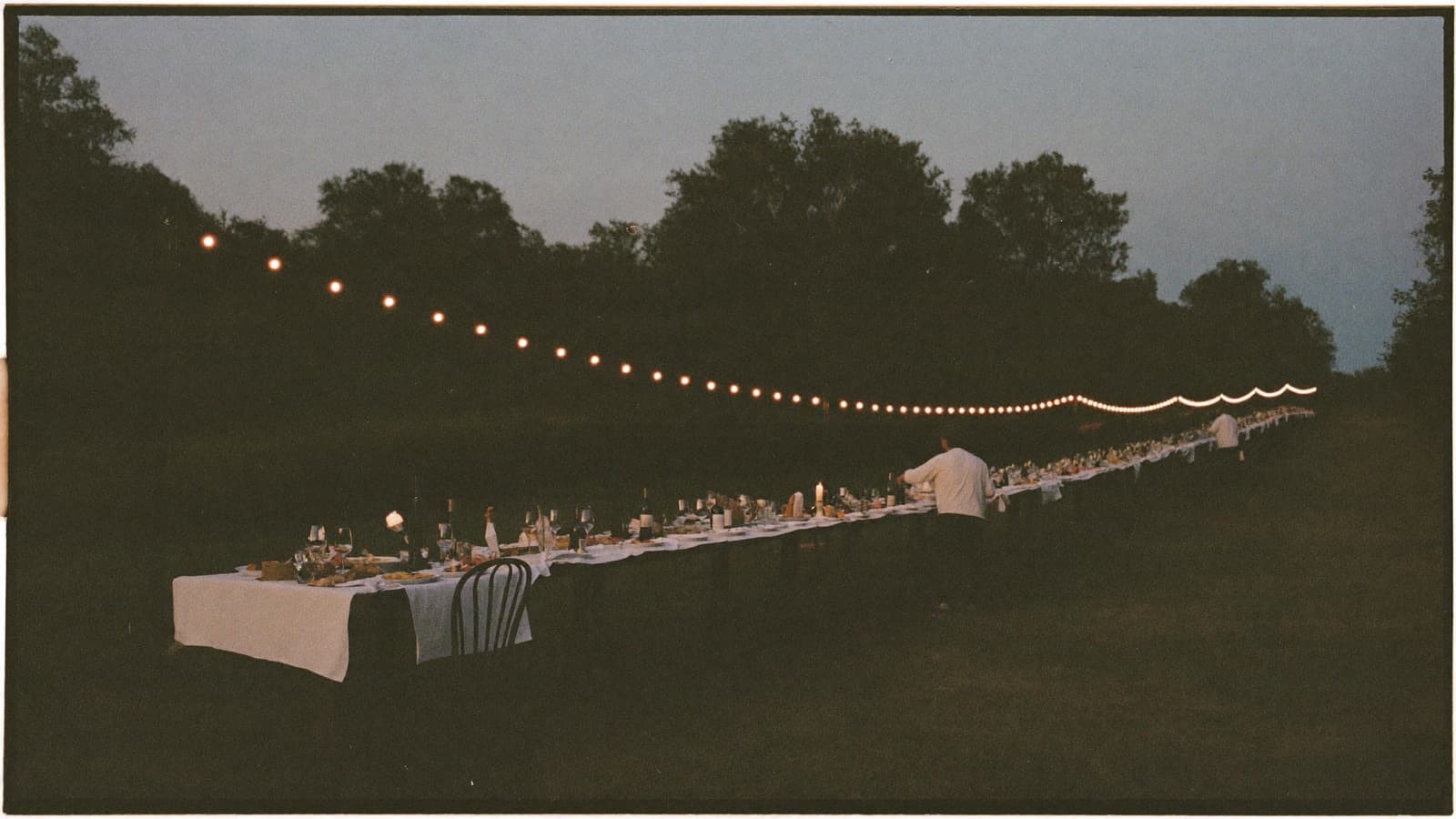 A crowd of friends at a long outdoor table at dusk, lit by string lights