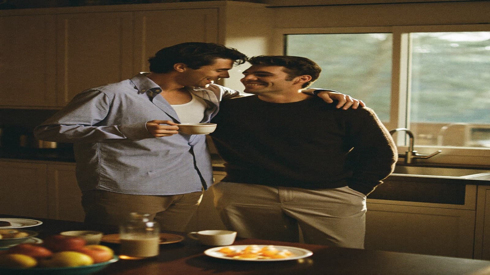 Two men laughing in a warm kitchen, one arm around the other's shoulder, breakfast on the counter