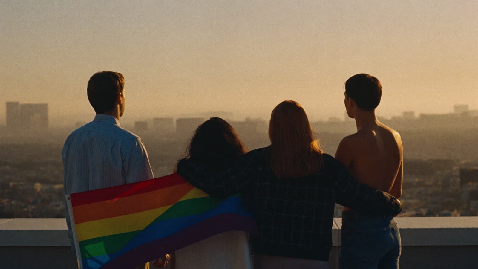 Four friends stand arm in arm on a rooftop at golden hour, one holding a rainbow flag, looking out over the city