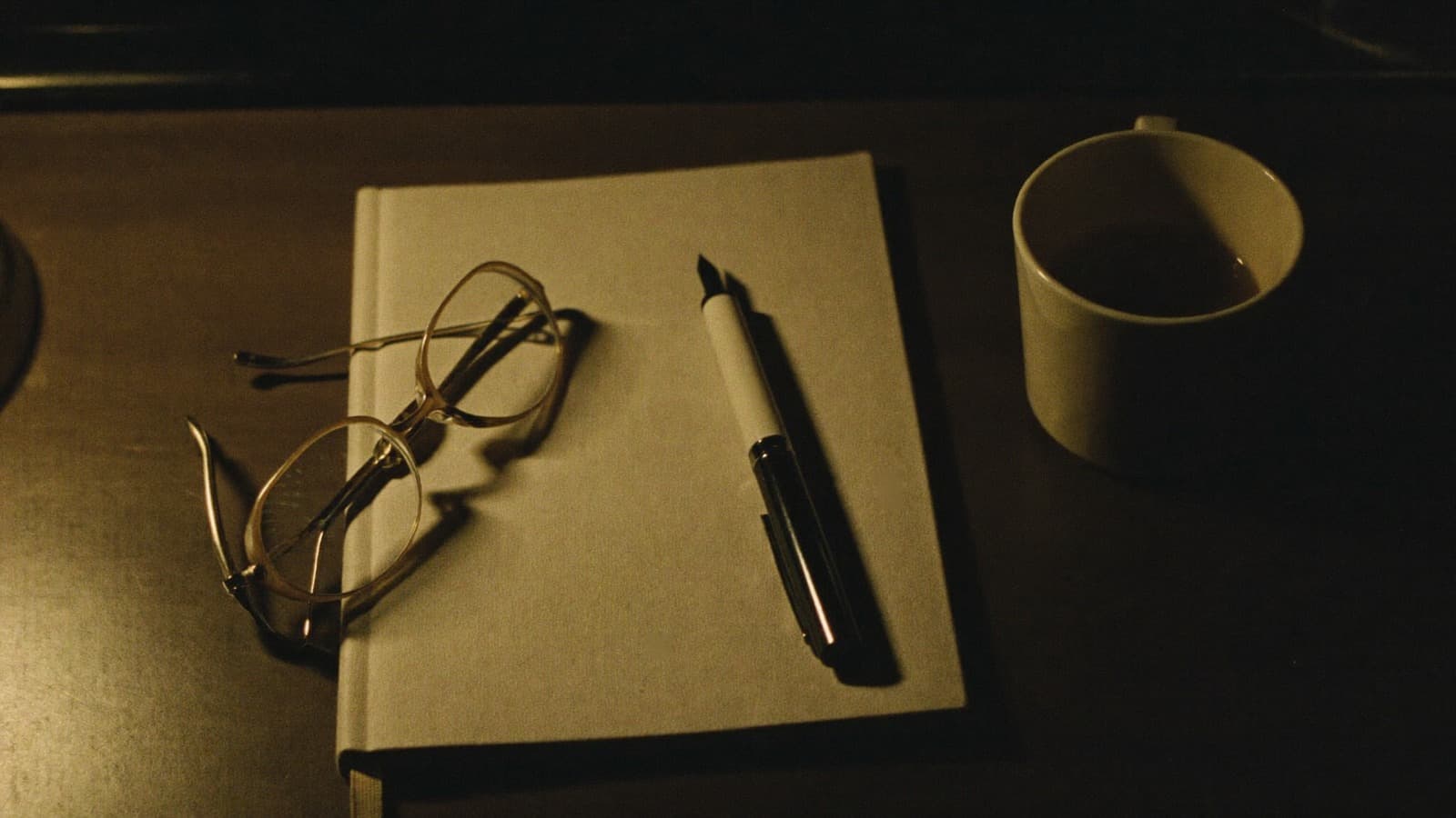 A closed notebook in lamplight, reading glasses and a fountain pen resting on it