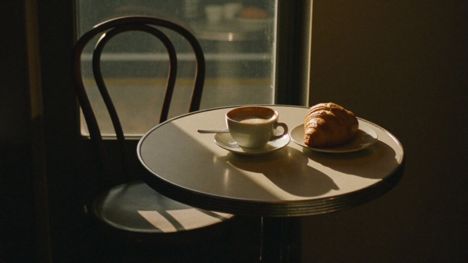 A coffee and a croissant on a café table by a window, an empty chair beside it